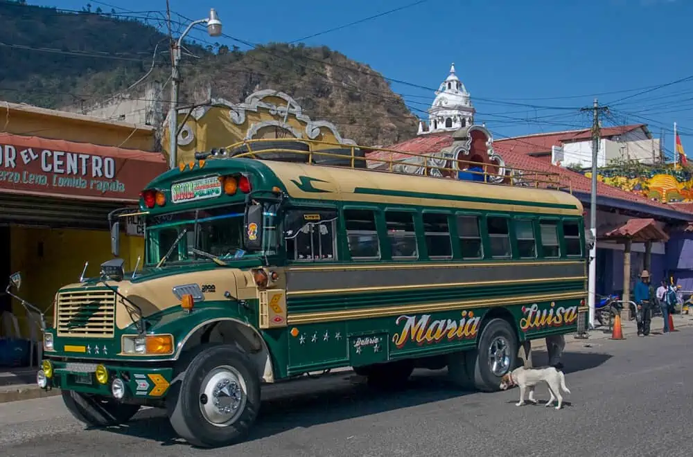 a chicken bus on the street in guatemala, a dog is standing next to it