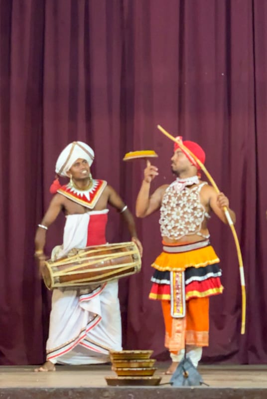 Two performers on stage, one playing a traditional drum and the other holding a bow, both in colorful costumes and headgear, engaging in a cultural dance or act among the things to do in Kandy