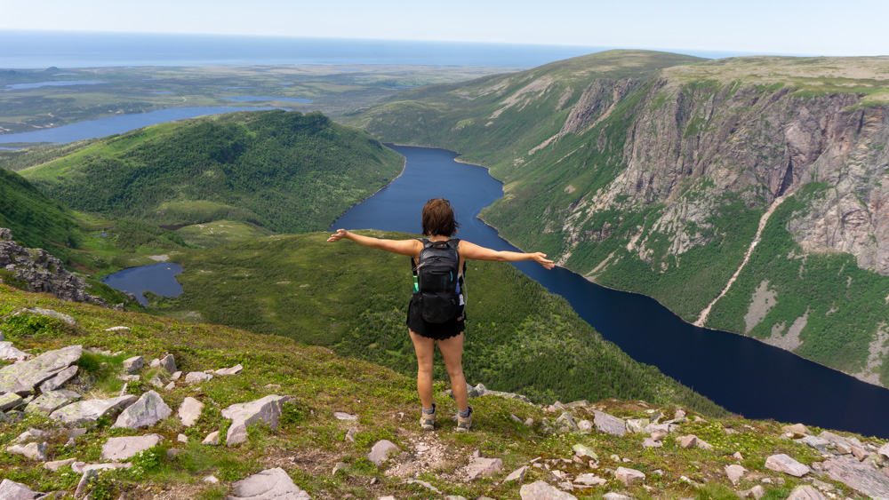 lora wearing a hiking backpack with arms wide open overlooking a beautiful view on top of gros morne mountain in newfoundland. in the background are winding rivers through the mountains.