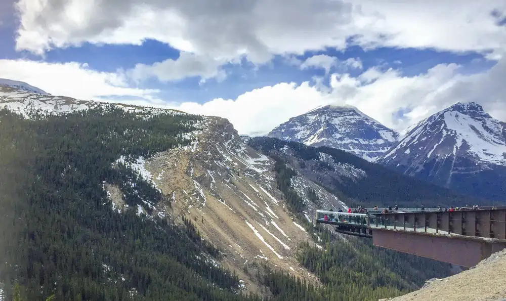 travelers standing on the skywalk in jasper national park. in the distance are large mountain peaks covered in snow.