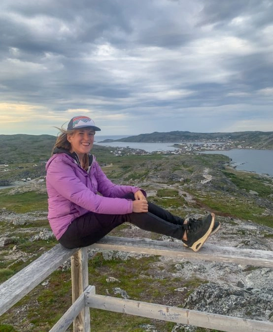 lora smiling while sitting on a ledge on brimstone head trail. behind her is the town of fogo.