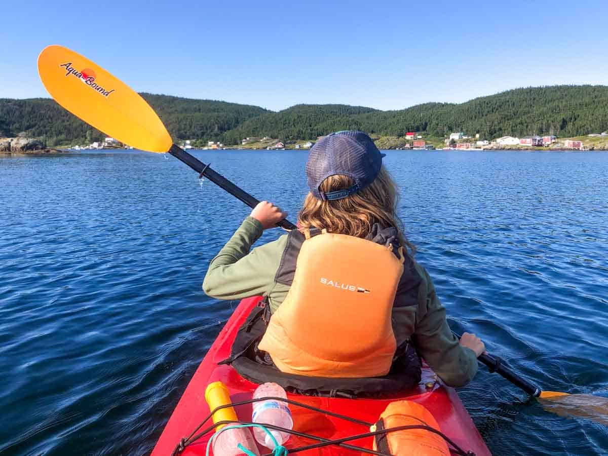 lora on a red kayak in newfoundland, she is wearing a life jacket and paddling through the scenic atlantic ocean with hills and colorful homes in the backdrop