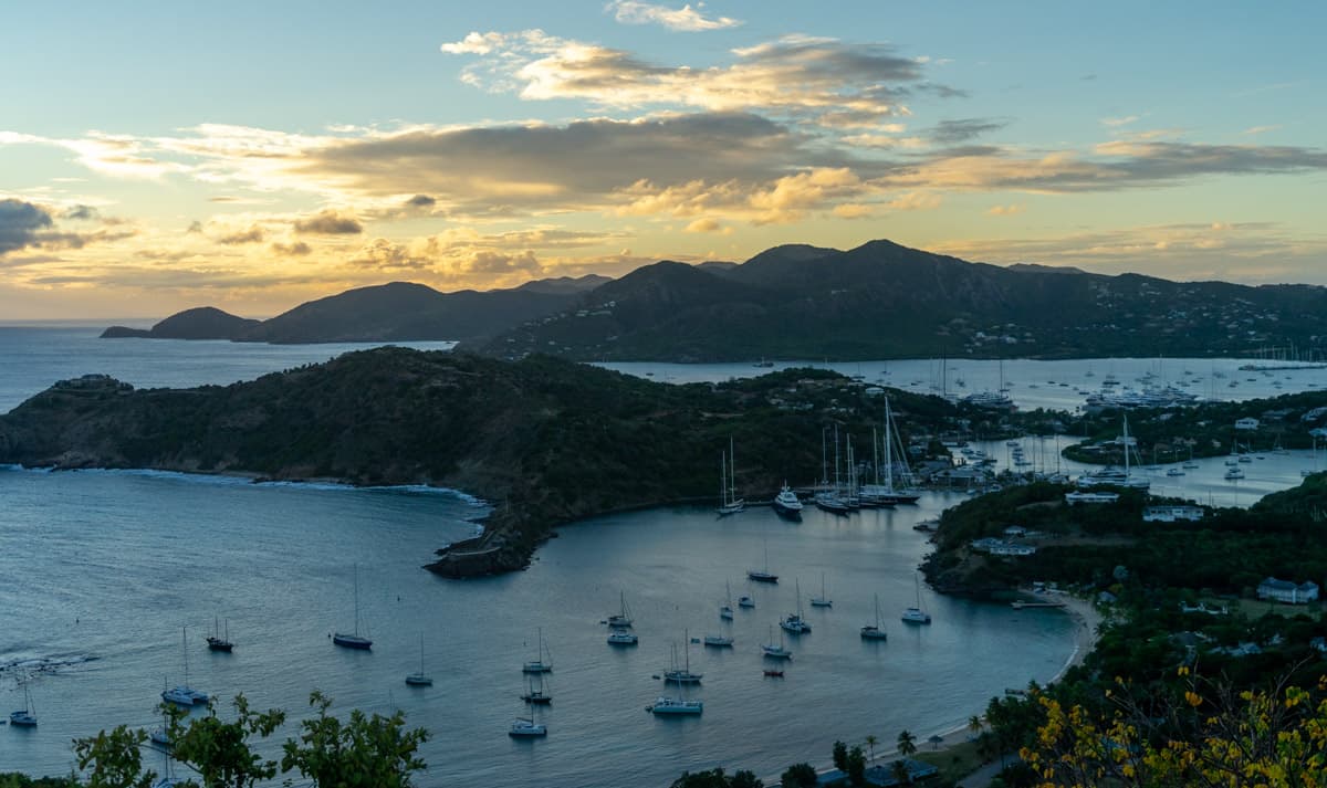 view of english harbour in antigua from shirley heights lookout oint. The sun is setting on the rolling hills and you can see dozens of sailboats in the habrour.