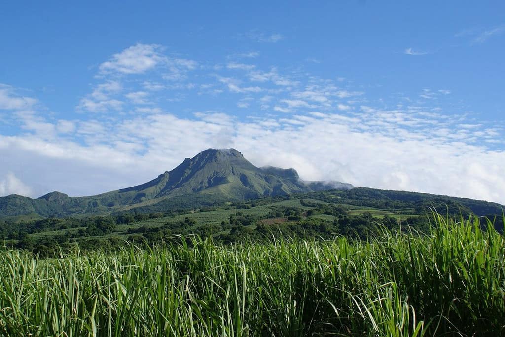 mountain in martinique centered in photo, green grass is in the foreground and the backdrop is a blue sky with clouds