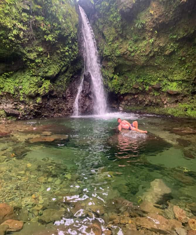 man lying on rock in emerald pool of water in front of jacko falls dominica
