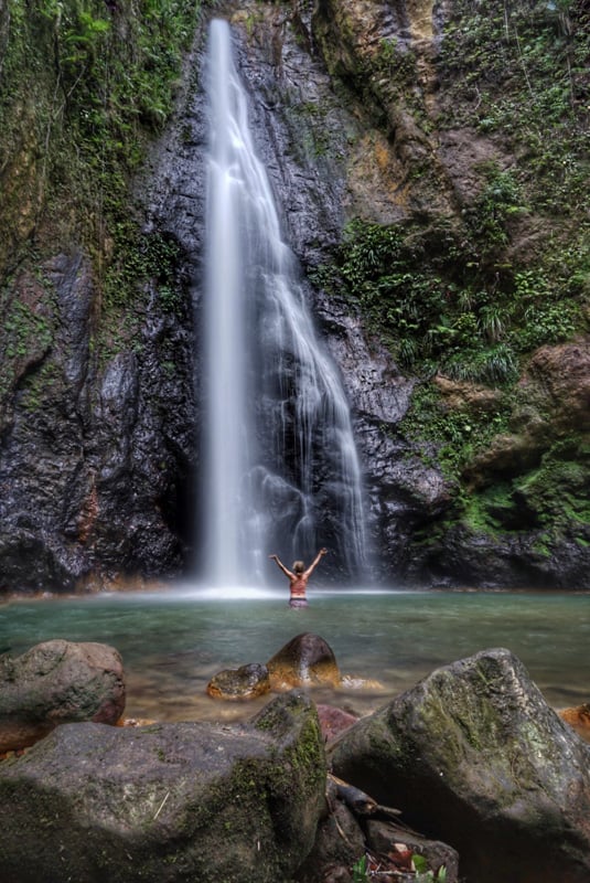 lora in front of syndicate falls on a hike in dominica. she is holding her hands up as the water comes pouring down.