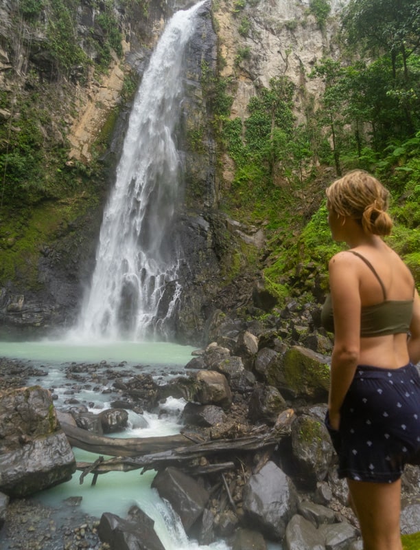 lora looking at the blue water of victoria falls while hiking in dominica. she is wearing shorts and a bikini top.