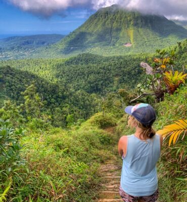 lora from the back on a hike in dominica. she is looking at a volcano in the distance with lush green tropical foliage around her.