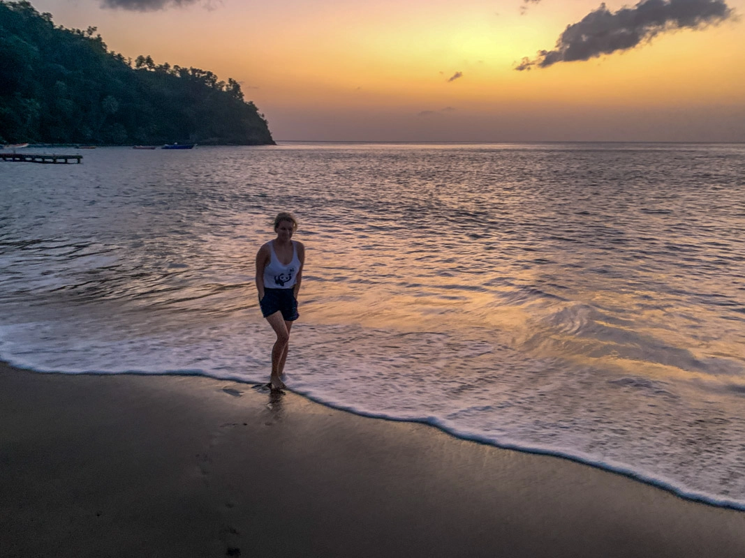 lora standing on toucari beaches in dominica. the waves are gently rolling in as the sun casts a purple and orange glow over the water.