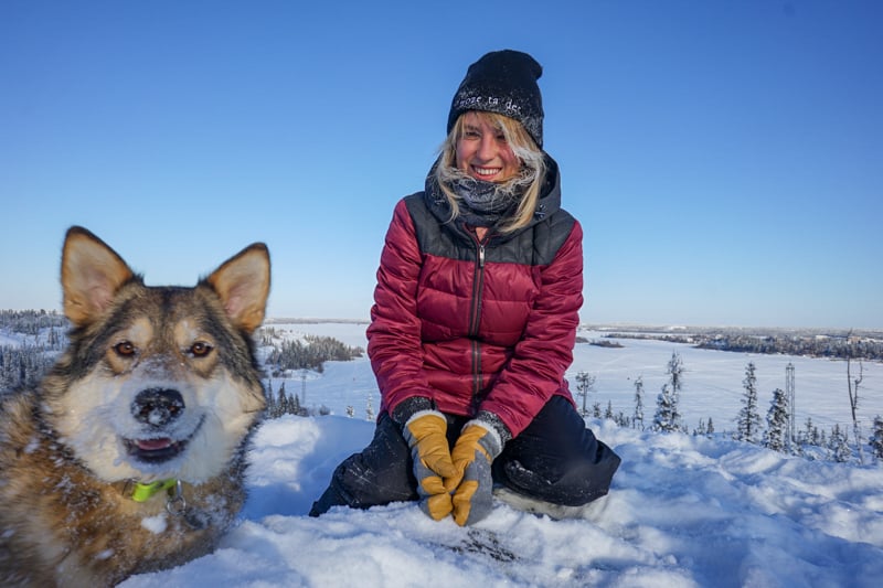 lora in yellowknife canada with a husky. she is wearing a winter jacket and gloves while playing in the snow. the dog has snow on its nose and is smiling at the camera.
