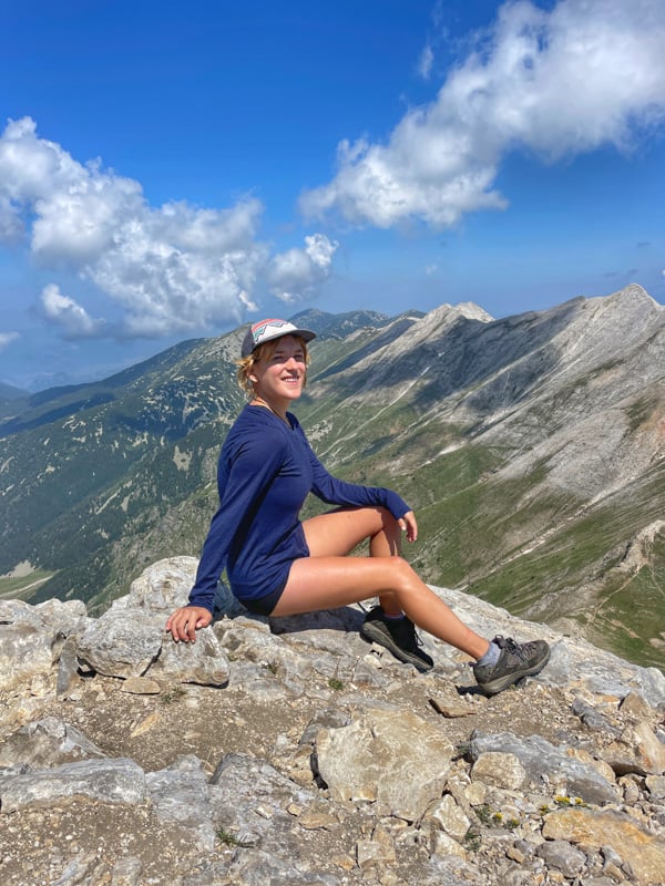 lora sitting on top of a mountain in bansko bulgaria after a hike. behind her are stunning mountain ranges. she is smiling looking proud.