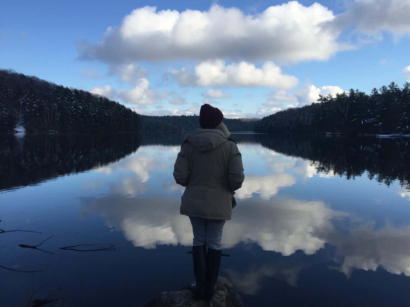girl wearing a winter jacket and hat staring out at a reflecting lake in muskoka. the clouds in the sky are reflecting in the water.