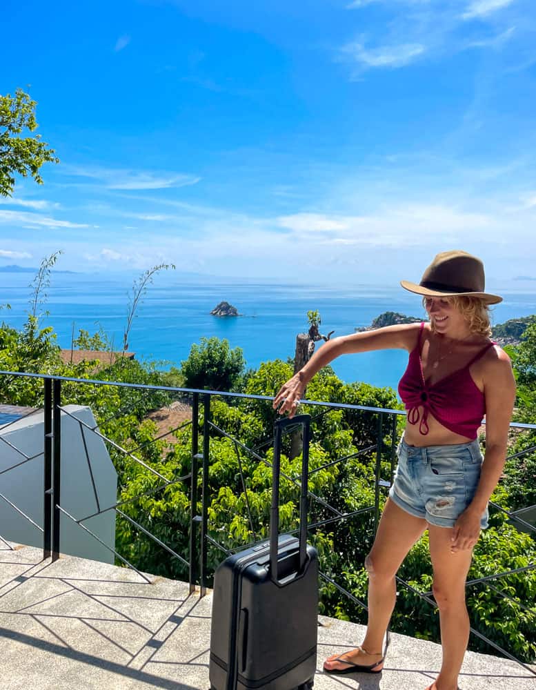 lora in thailand with nomatic carry-on suitcase, behind her is the ocean.