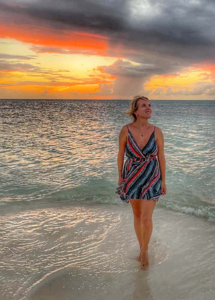 lora wearing a red and blue dress standing bearfoot in the water as the sun sets behind her in shades of red and orange