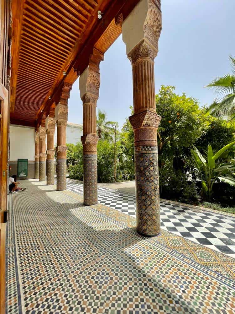 architecture in marrakesh morocco. 5 tall pillars painted in geometrical designs next to a black and white tiled floor. plants surround the building.