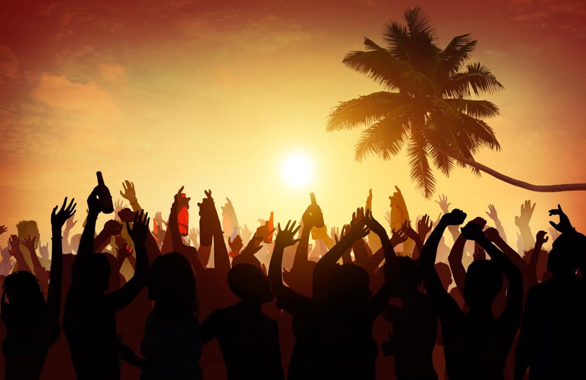 A silhouette of a lively crowd with raised hands against a sunset with palm trees in the background, capturing the essence of party cities in Mexico.