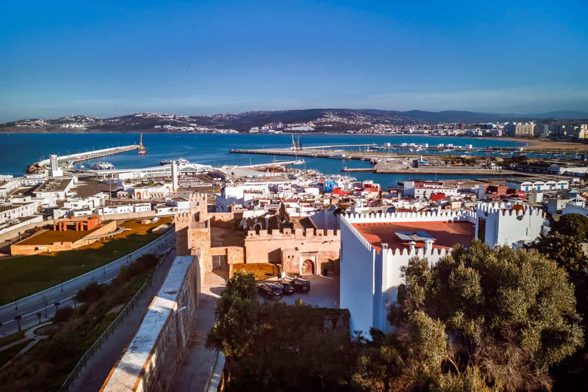The old medina and the port of Tangier, Morocco. Photo shows buildings with ocean and mountains in backdrop.