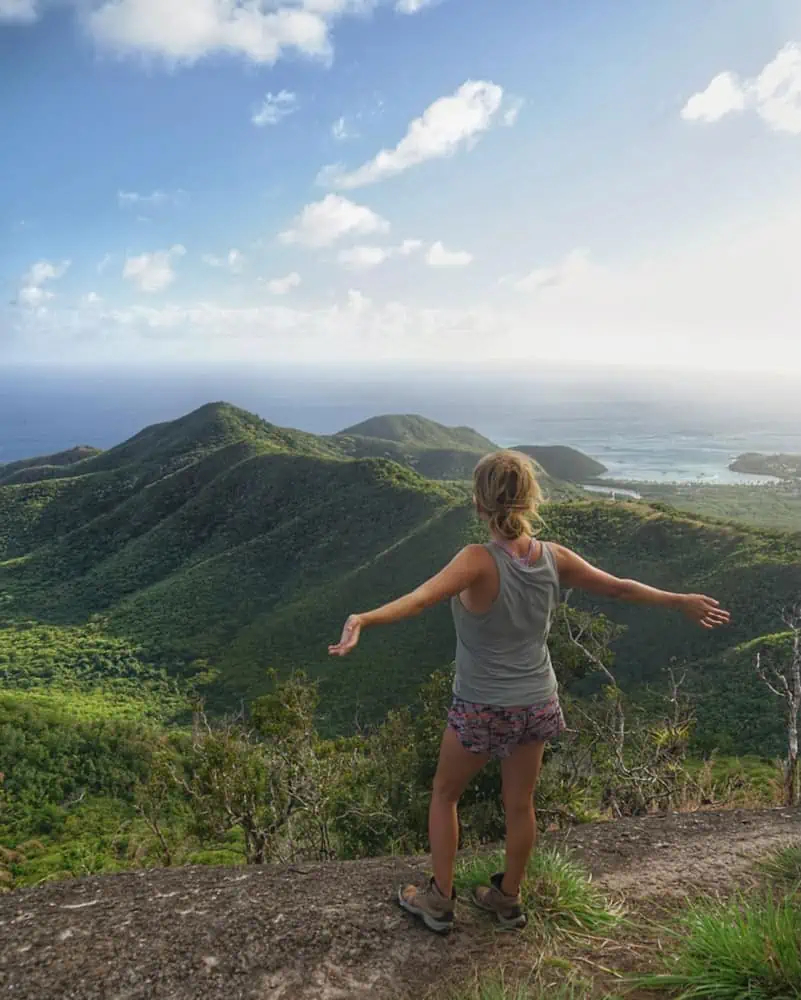 lora from the back with arms out wide overlooking rolling green hills with the blue ocean behind them 