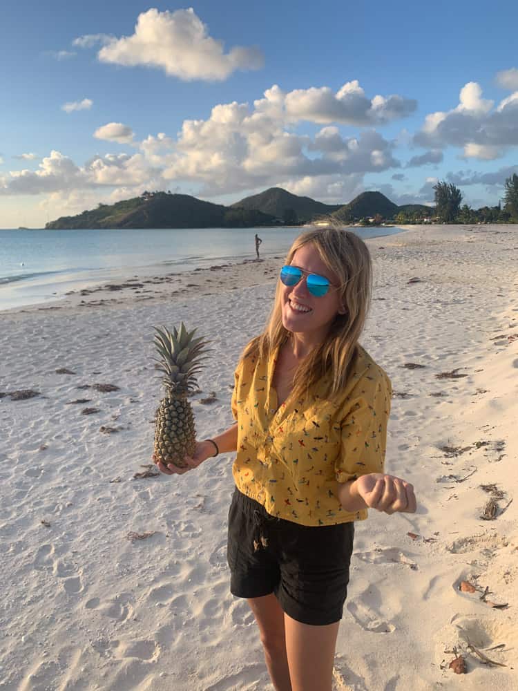 lora holding a tiny pinnaple in Antigua on the beach. she is wearing a yellow t shirt and blue glasses, smiling at the camera.