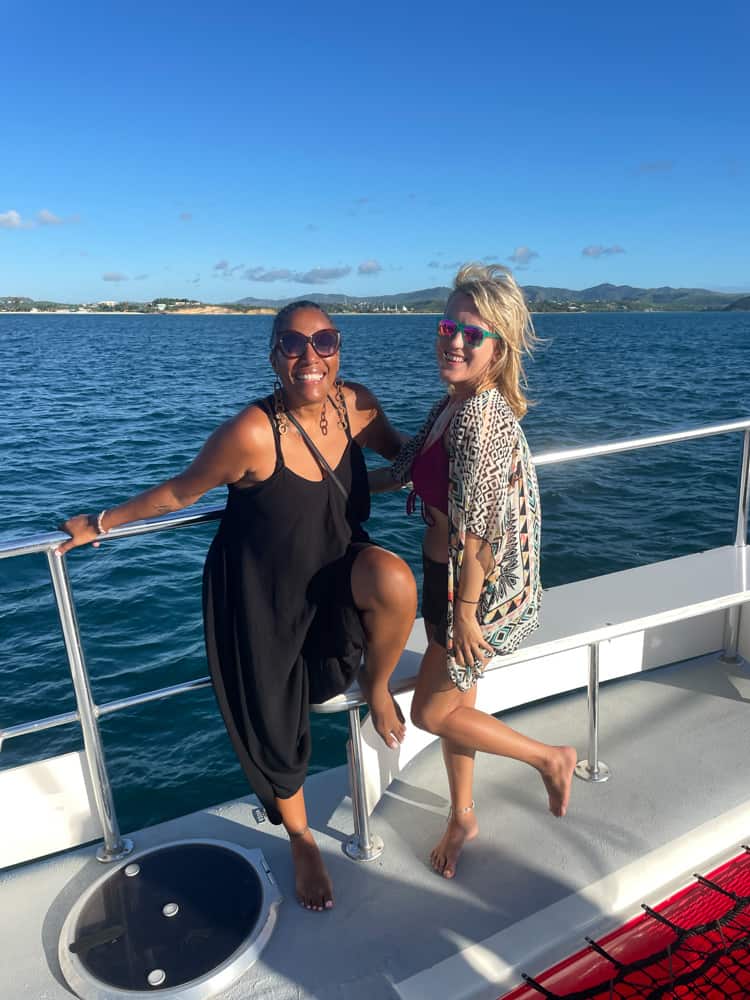 lora and friend smiling looking at the camera on a catamaran in antigua. the ocean and hills are the backdrop.