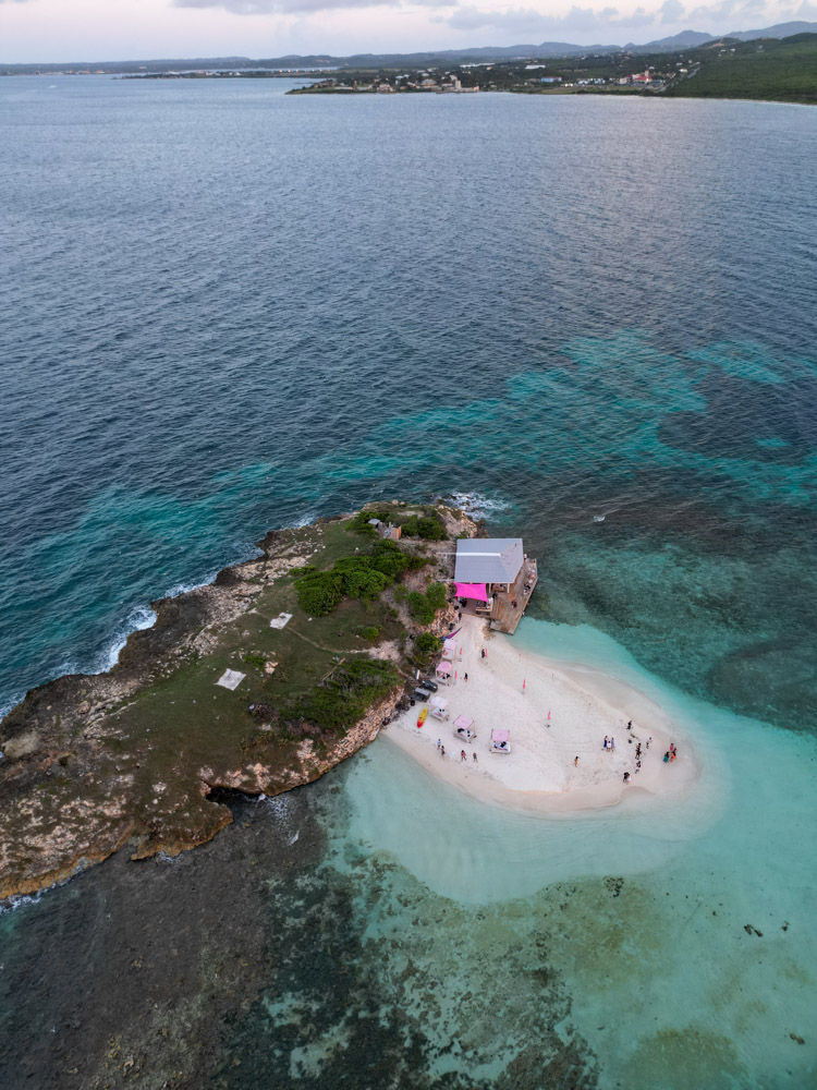 aerial view of prickly pear island in antigua. travelers stand on the white sand island with shades of blue water around it.