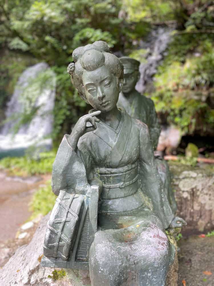 A statue of a japanese woman sitting near a waterfall.