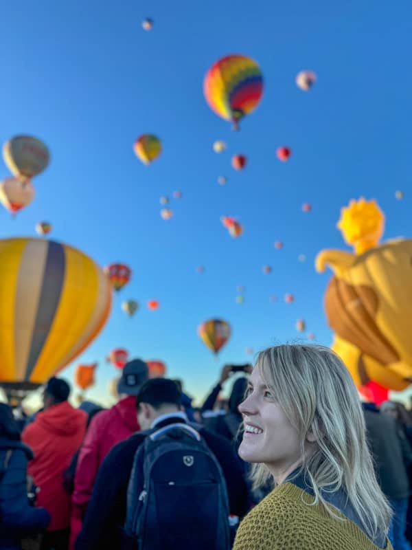 Woman smiling and looking up at numerous colorful hot air balloons in a clear blue sky during a balloon festival.