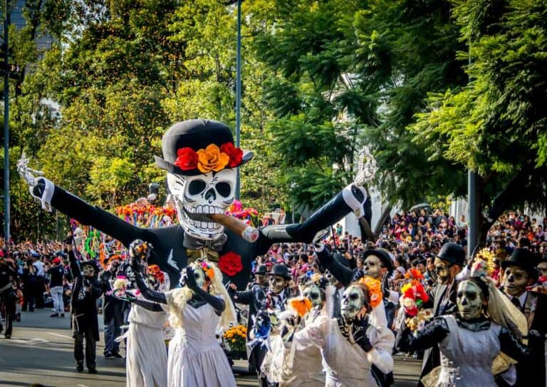 A vibrant day of the dead parade with participants in skeleton costumes and makeup, featuring a large puppet of a smiling skull adorned with flowers.