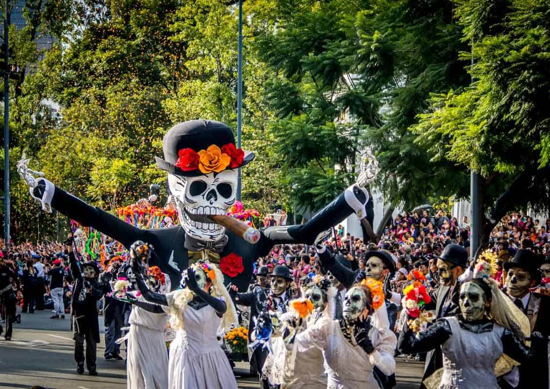 A vibrant day of the dead parade with participants in skeleton costumes and makeup, featuring a large puppet of a smiling skull adorned with flowers.