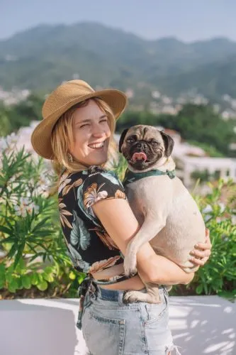 A woman in a hat smiles while holding a pug outdoors, with mountains and greenery visible in the background.