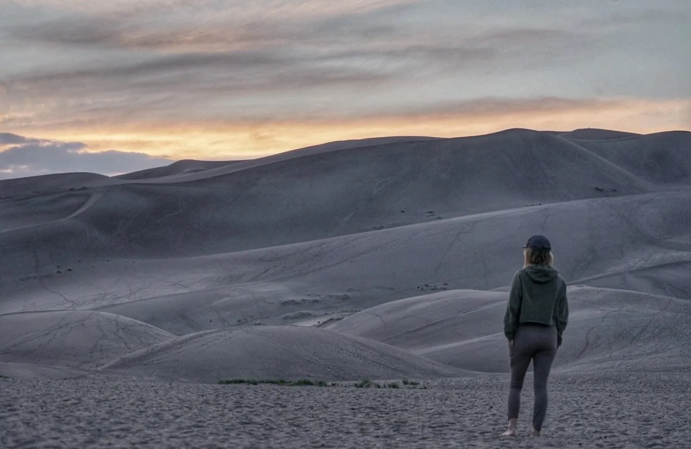 A person in casual clothing stands on sandy ground, looking at large, rolling sand dunes under a cloudy sunset sky.