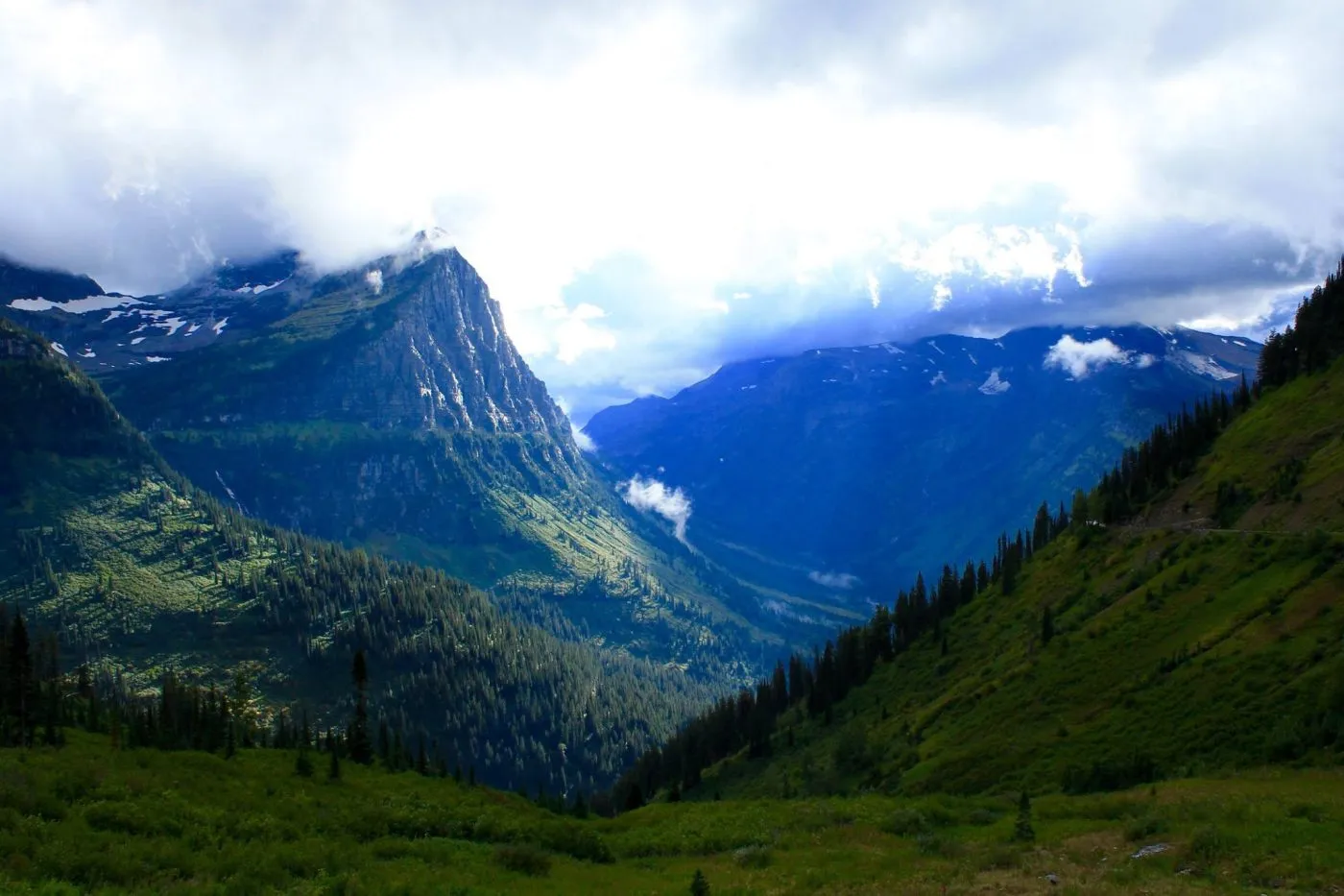 A lush green valley with steep, forested mountains under a cloudy sky, sunlight illuminating parts of the landscape.