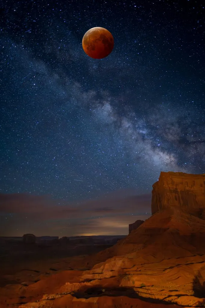 A reddish moon is visible above a desert landscape at night, with the Milky Way stretching across a clear, star-filled sky.