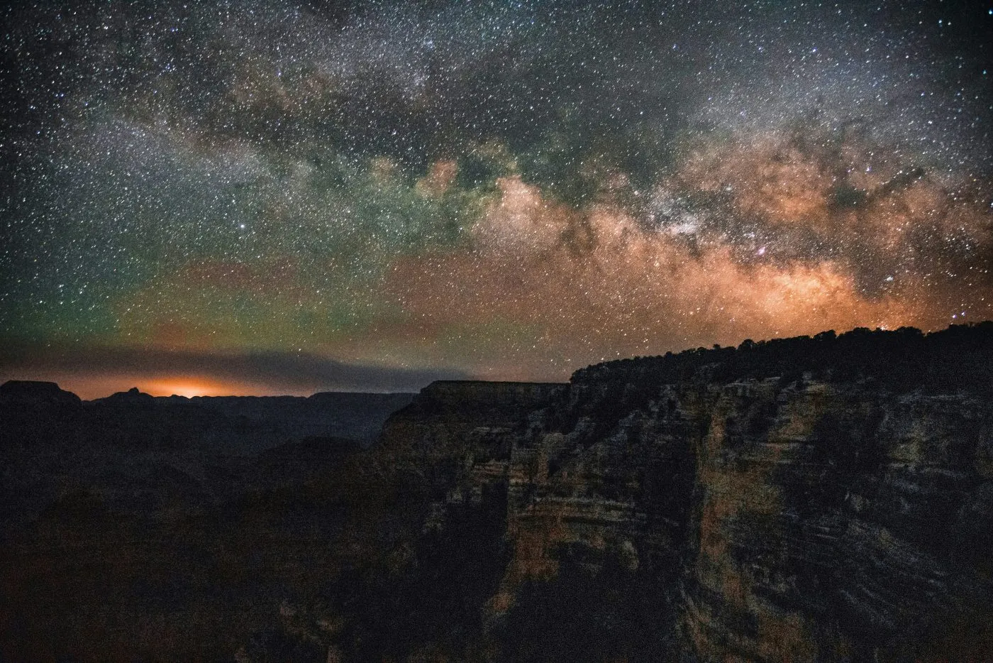The Milky Way galaxy glows brightly in the night sky above rocky canyon cliffs, with a faint orange light on the horizon.