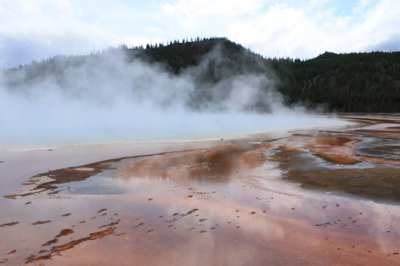 Steam rises from a colorful geothermal hot spring with mineral terraces, set against a backdrop of forested hills and a cloudy sky.
