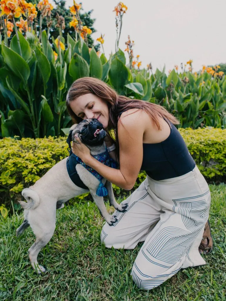 Woman kneeling on grass hugs a pug wearing a blue harness in a garden with orange flowers and green foliage in the background.